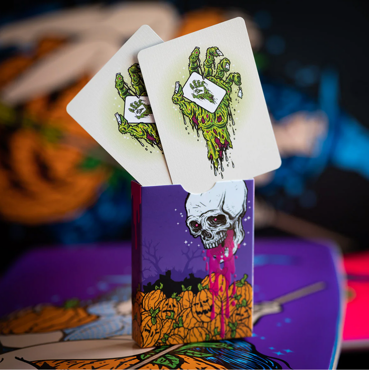 Two playing cards with green zombie hands on white backgrounds, held above a purple card with a skull and pumpkins design.