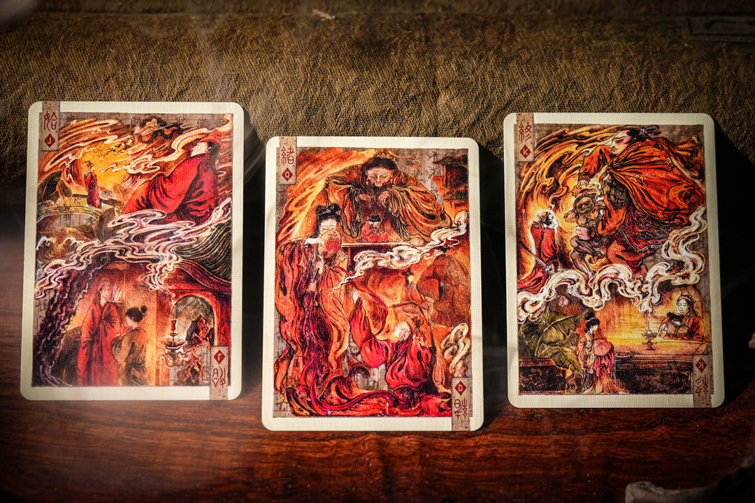 Three illustrated tarot-style cards are arranged upright on a dark wooden surface with a textured brown background. The card art is dominated by fiery red, orange, and yellow tones with dark shadowed areas, and no visible brand text is legible.