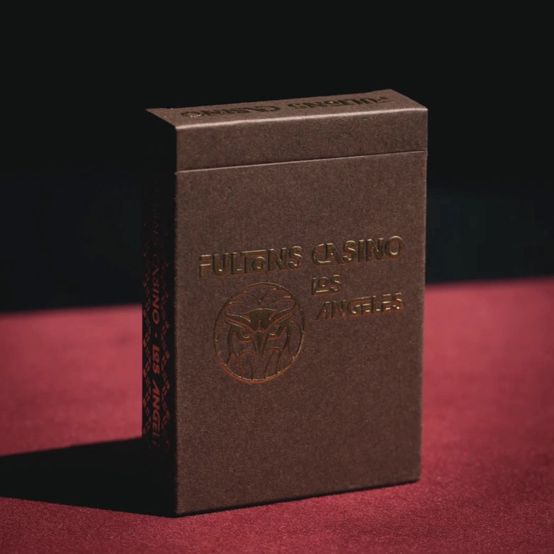 A dark brown playing card deck box with embossed gold lettering reading 'FULTON'S CASINO LAS ANGELES' and a circular fox logo, standing on a red surface against a dark background.