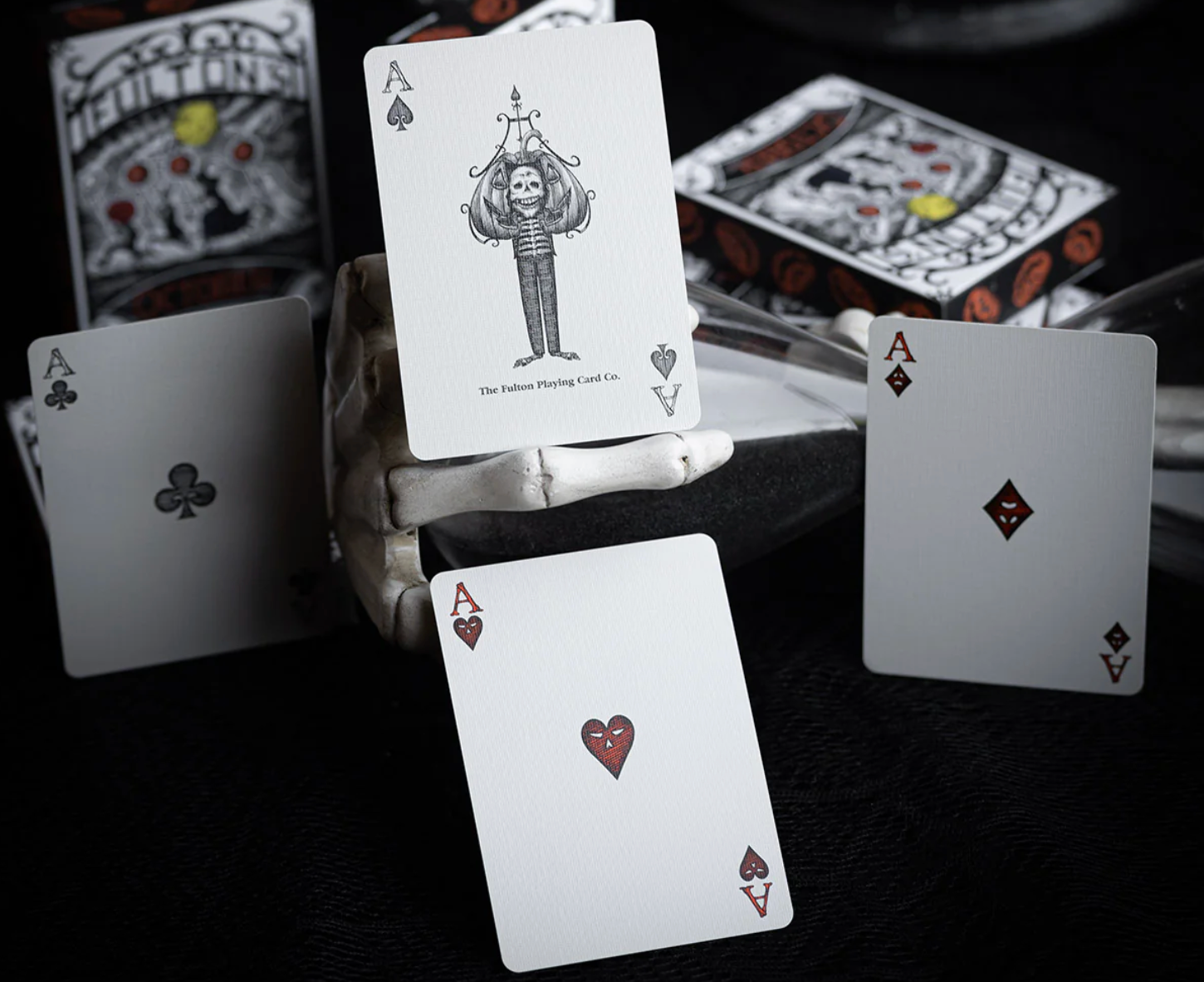 Four aces from a unique deck of cards on a dark surface with additional cards and a box in the background.