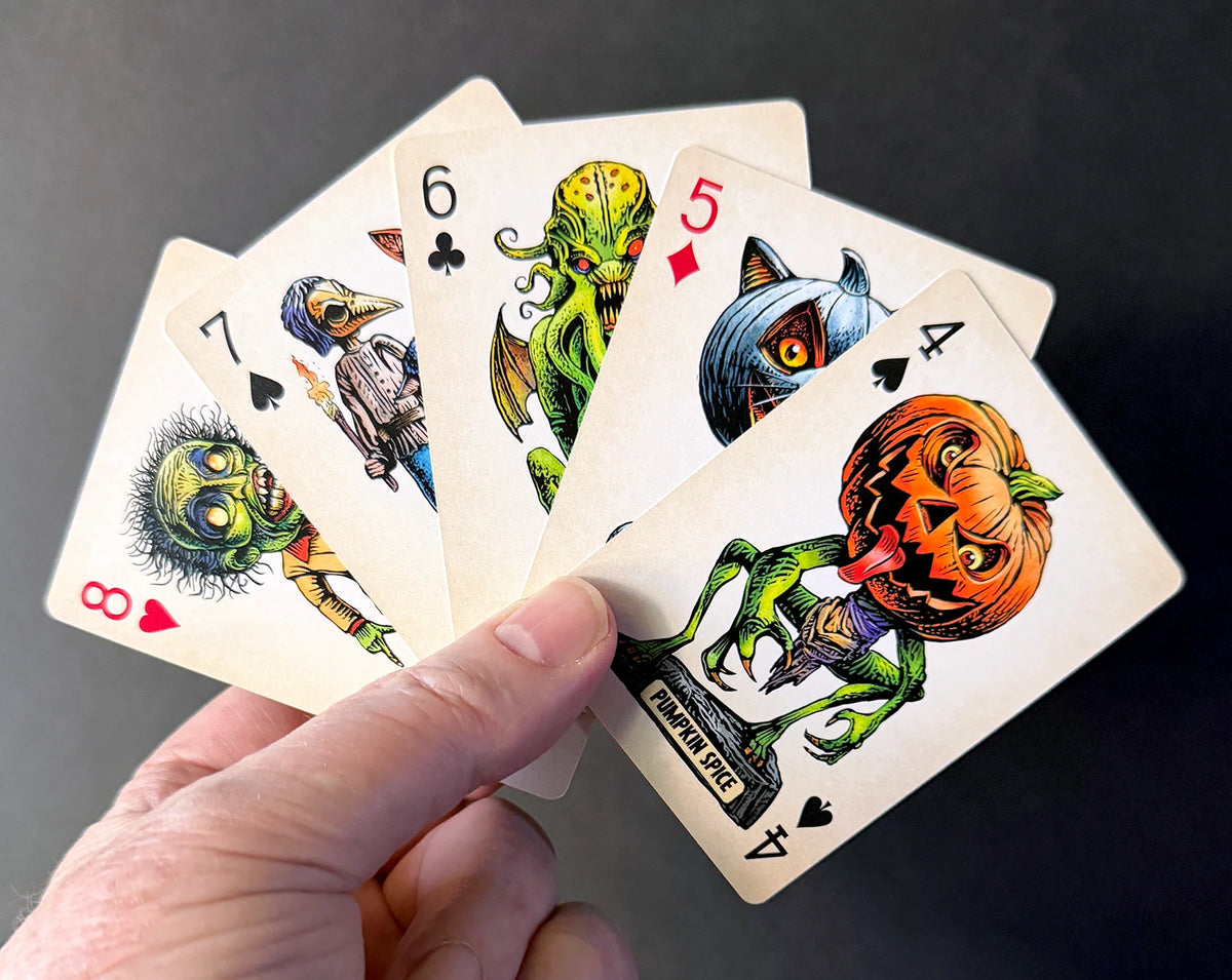 A hand holds a fan of five illustrated playing cards with Halloween-themed designs, including a pumpkin-headed figure, a cat-headed card, and zombie-like cards, against a dark background.