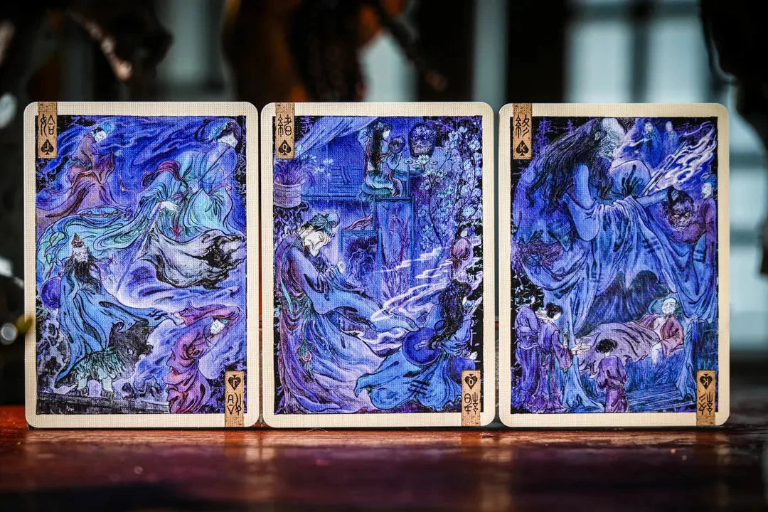 Three illustrated playing cards with swirling blue and purple artwork are displayed upright on a wooden surface, framed by a dark, blurred background.
