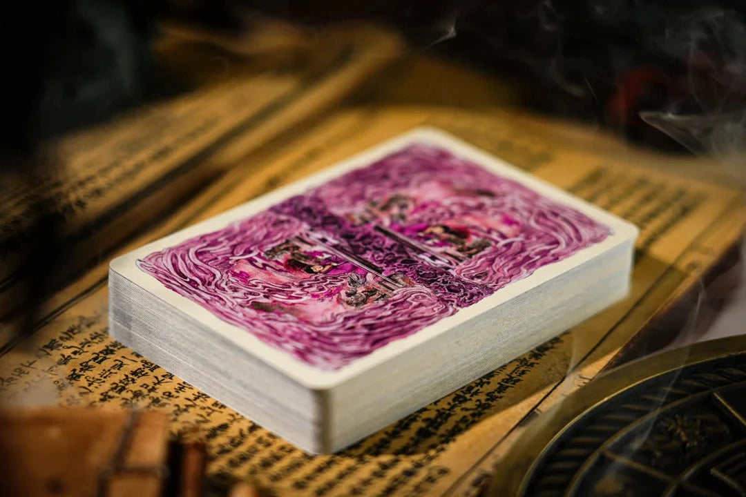 A deck of playing cards with a purple and pink abstract pattern on the back, resting on an old book surrounded by smoky, mystical props.