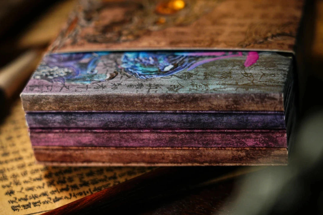 A close-up of a decorative stack of cards or a deck box with a fantasy illustration on the top, resting on an old open book. The product has a weathered brown base with purple, blue, and pink illustrated accents.