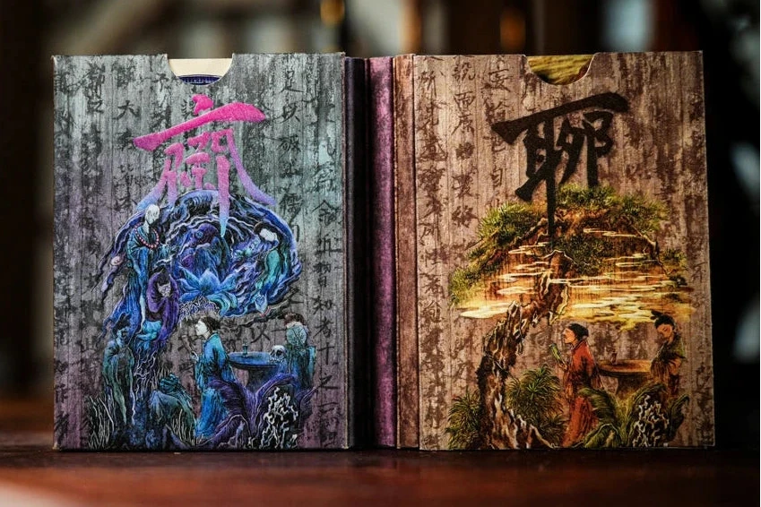 LiaoZhai Playing Cards decorative book style playing card boxes standing upright on a wooden surface, featuring detailed illustrated covers with Chinese characters and fantasy-style artwork in blue, purple, and warm brown tones