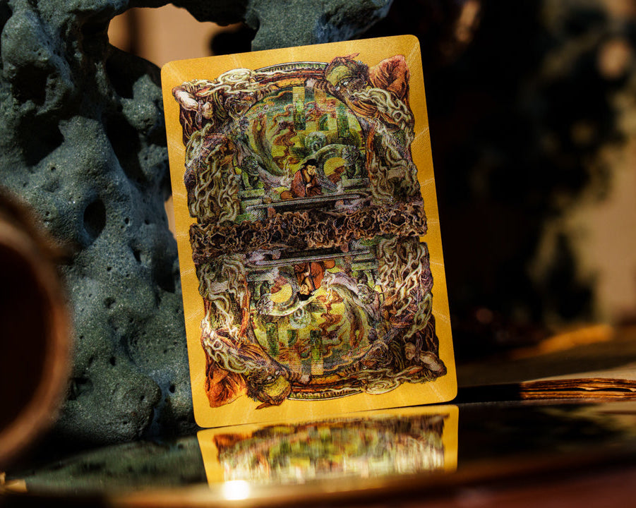 A single illustrated playing card with a yellow border and dense fantasy artwork stands upright on a reflective surface, surrounded by dark decorative props in a moody indoor setting.