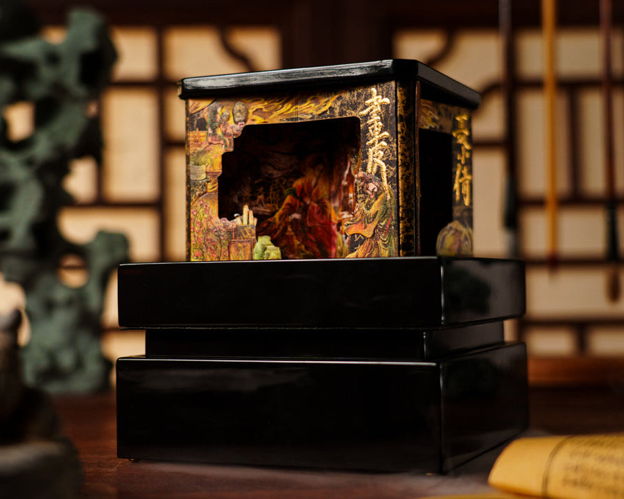 A small black lacquered box with a decorative gold and multicolor illustrated lid, displayed on a wooden surface with a blurred traditional interior background.