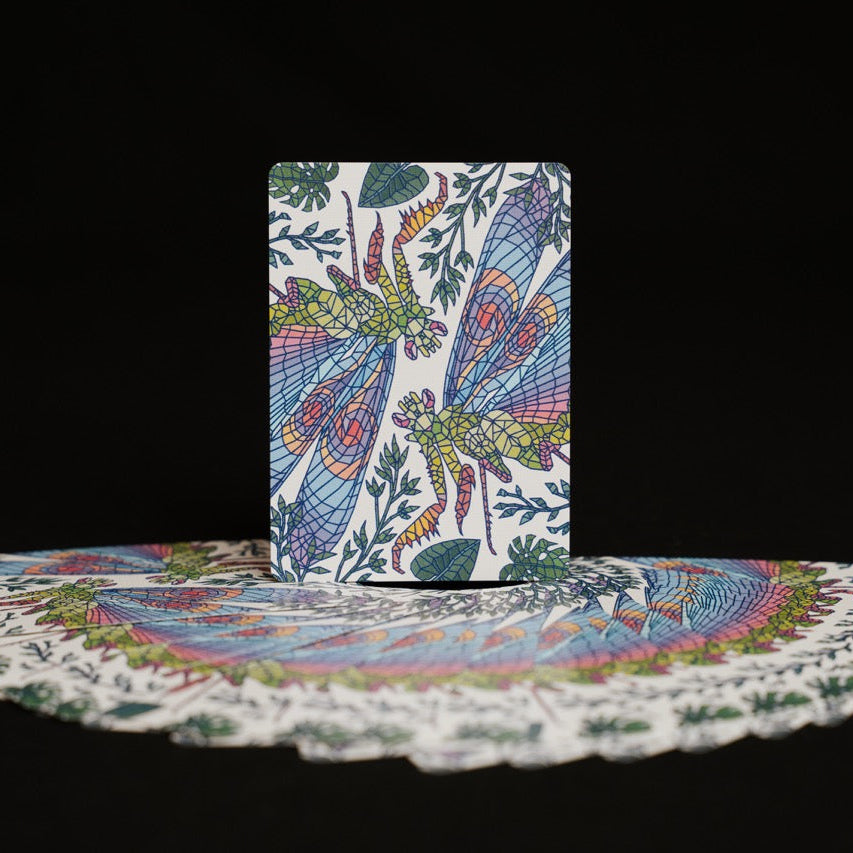 A deck of illustrated cards standing upright with several cards fanned out in front, all showing a detailed multicolor dragonfly pattern on a white background, set against a solid black backdrop.