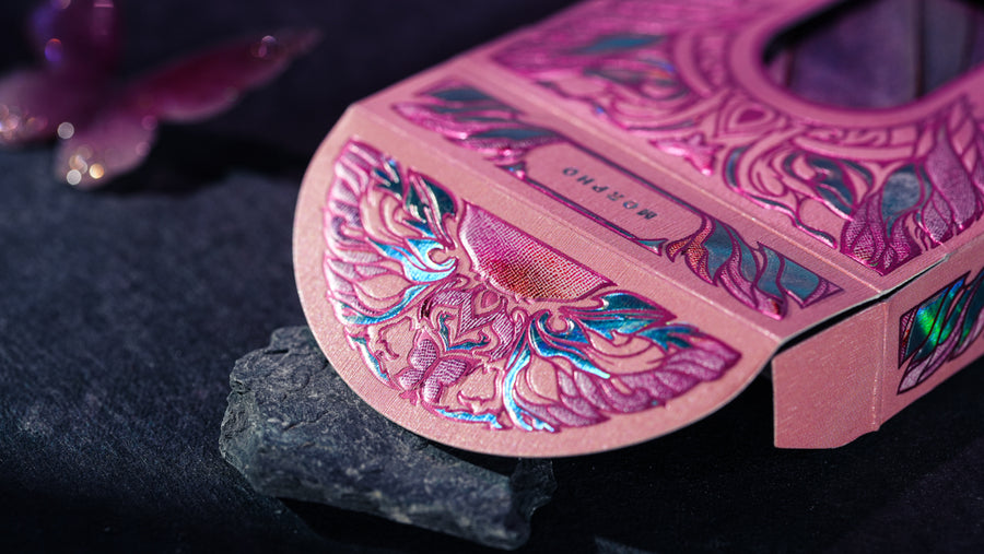 A close-up of a pink decorative tuck box with intricate metallic foil patterns in blue, green, and silver, resting on a dark textured surface.