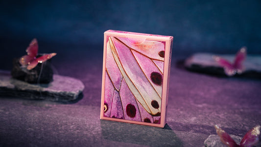A pink playing card deck box standing upright on a dark textured surface, featuring a butterfly wing pattern in shades of pink, purple, and cream with dark spots.