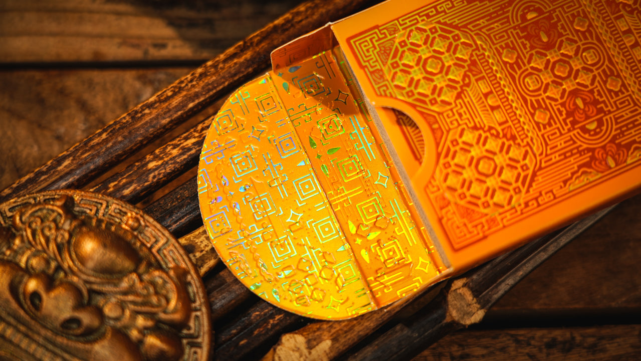 A close-up of an ornate orange and gold playing card deck with a matching box, partially open to reveal a circular card back decorated with holographic Chinese characters, resting on a rustic wooden surface.