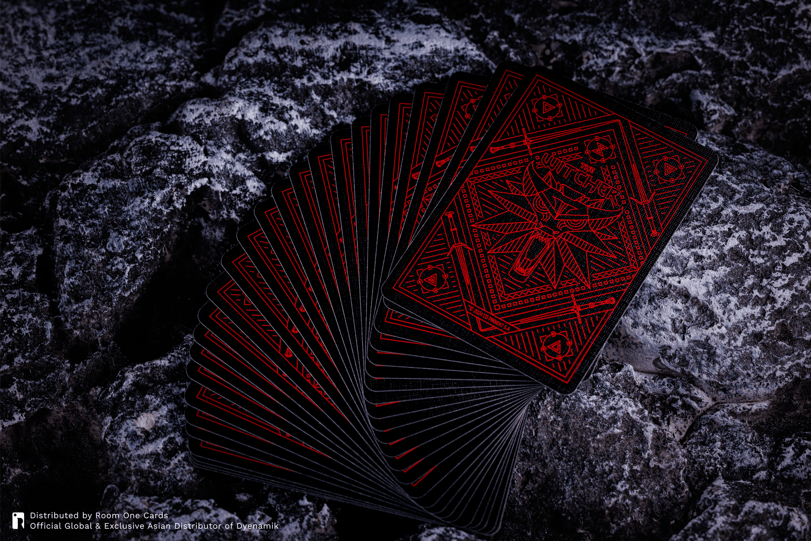 A fan of black playing cards with intricate red occult-style geometric designs, displayed on a dark, textured rock surface. The Witcher Playing Cards by Room One.