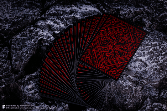 A fan of black playing cards with intricate red occult-style geometric designs, displayed on a dark, textured rock surface. The Witcher Playing Cards by Room One.