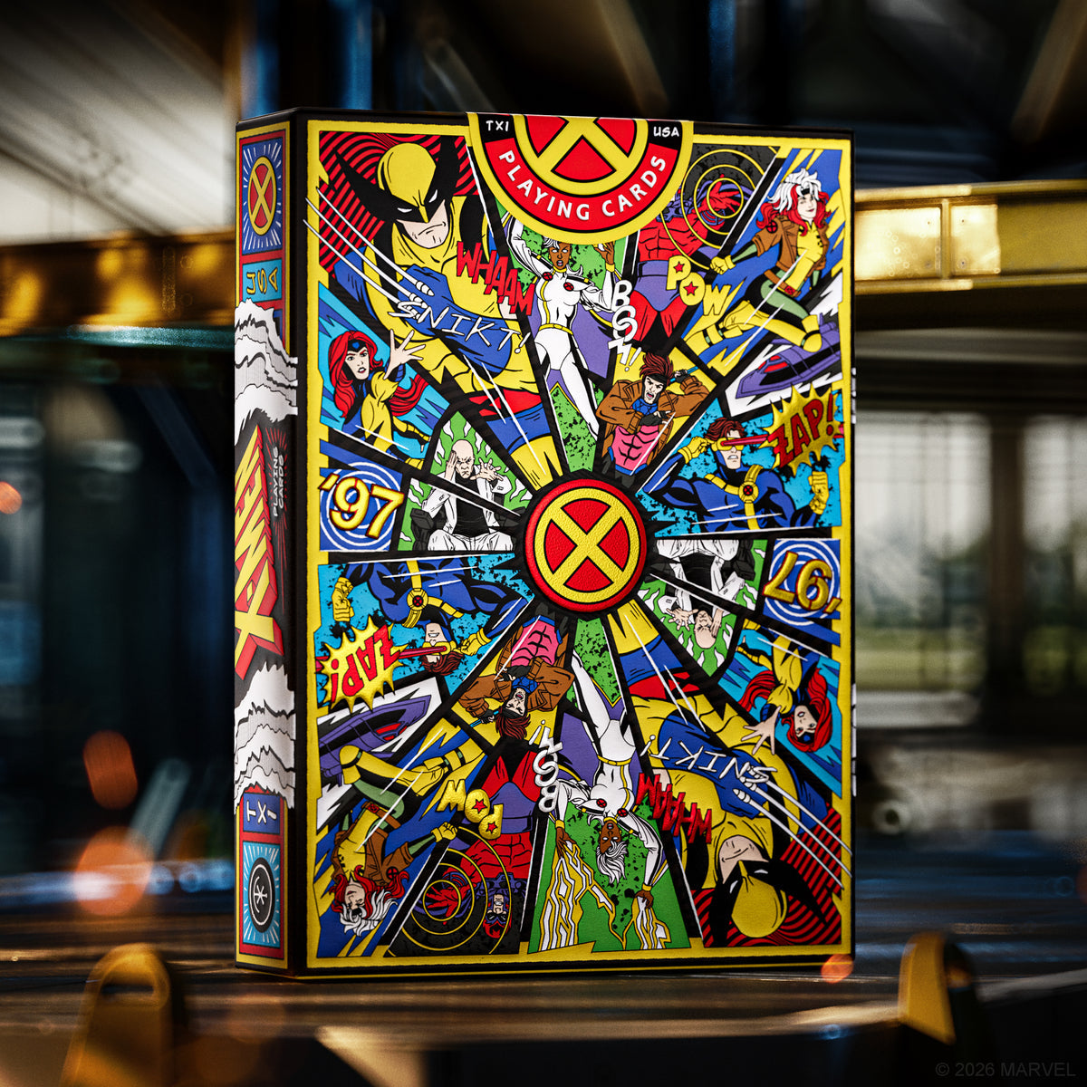 A colorful comic-book style playing cards box featuring X-Men characters and artwork, photographed upright in a blurred indoor setting with warm ambient lighting.